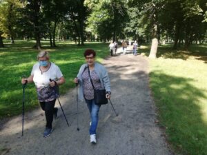2 idące osoby z kijkami, za nimi w oddali kolejne osoby na spacerze w parku - zajęcia taneczno-ruchowe w Klubie Seniora