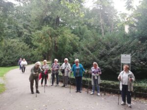 stojąca grupa osób z kijkami na tle drzew - zajęcia taneczno-ruchowe w Klubie Seniora