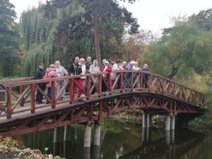grupa osób spaceruje z kijkami na łonie natury, stojąca na moście - zajęcia taneczno-ruchowe w Klubie Seniora