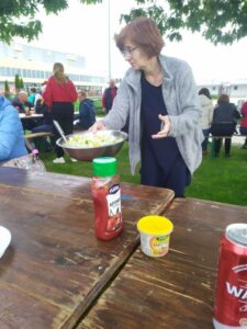 kobieta trzyma w ręku miską z sałatką, ludzie na zewnątrz przy stołach