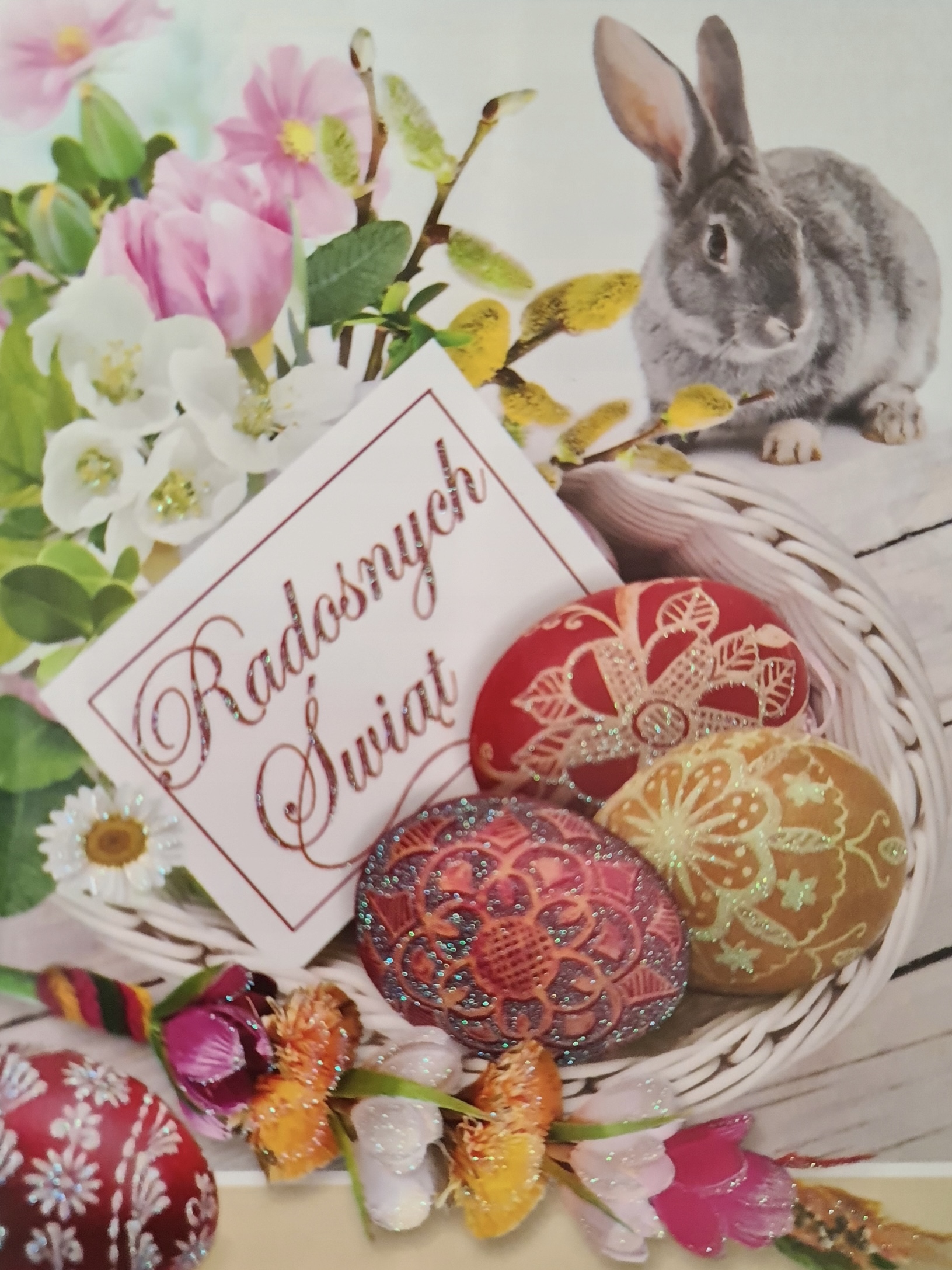 kartka świąteczno-wielkanocna. W koszyczku ozdobione jajka z włożoną kartką z napisem Radosnych Świąt. Koszyczek leży na stole, obok koszyczka siedzi zajączek. W prawym górnym rogu kwiatki białe i różowe