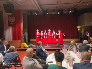 6 kobiet ubranych w czerwone suknie z białymi szalami na scenie śpiewa