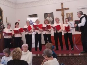 7 kobiet śpiewa z pozycji stojącej, w ręku trzymają czerwone śwpiewniki, po prawej mężczyzna gra na akordeonie