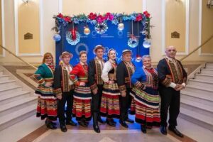 Osoby przebrane w stroje ludowe, zdjęcie wewnątrz pomieszczenia na tle ozdobionej ściany