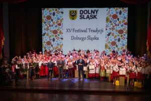 scena pełna ludzi ubranych w stroje ludowe, kilka osób jest w garniturach, w tle wyświetlony slajd: XV festiwal Dolnego Śląska